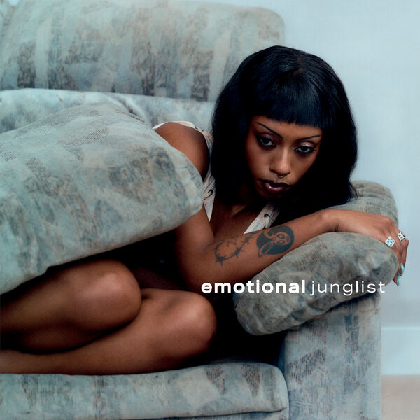 A Black woman, curled up on a sofa; the grey sofa cushion is covering most of her body like a comfort blanket. She looks to be in a great deal of emotional turmoil.