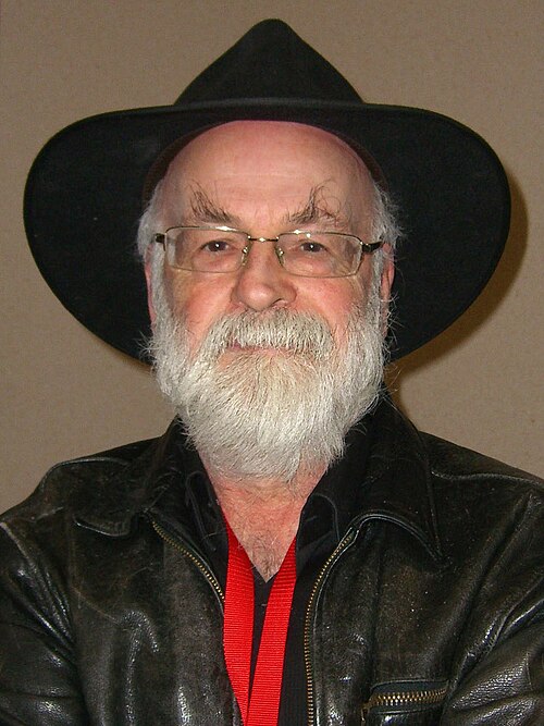 A photo of a white man. He has a short white beard and is wearing glasses and a wide-brimmed hat.