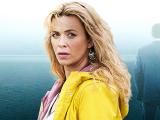 A woman looking pensively towards the camera. She is wearing a yellow coat, and her long hair falls down her back. Behind is a silhouette of a man facing away, and the sea is in the background.