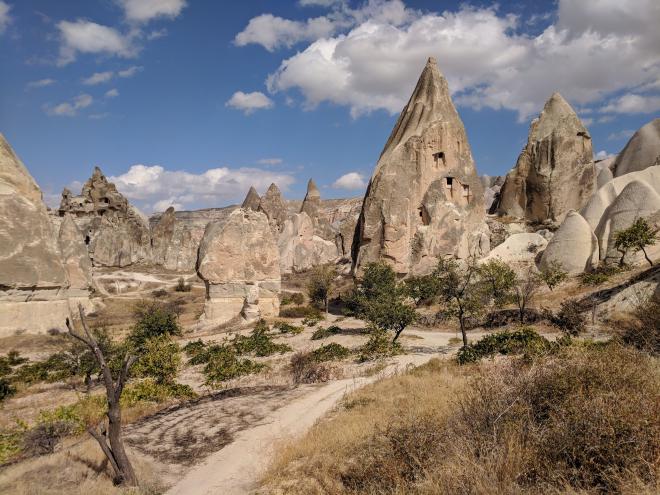 A view of some craggy rock formations. There are a number of different types: flat-faced, squat, square ones; tall, angular, smoother ones; some jagged and foreboding; others rounded and smooth; and a few more like rolling dunes. Some of the larger formations have windows and doorways carved into the side, hinting at some former habitation. In the distance the horizon is dominated by what appear to more cliffs. The foreground is arid, dry, but dotted with green bushes and a few spindly green trees. There is a dry dirt path leading into the rock formations.